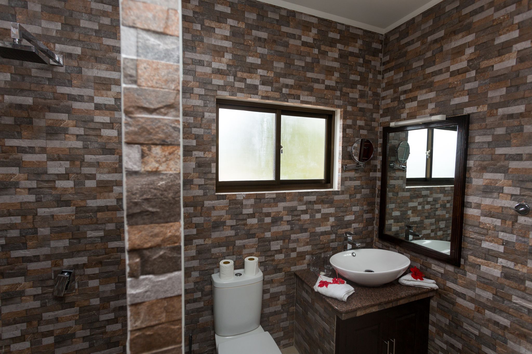 Un baño con pared de diseño de piedra, un inodoro, un lavabo con espejo y una ventana sobre el inodoro.