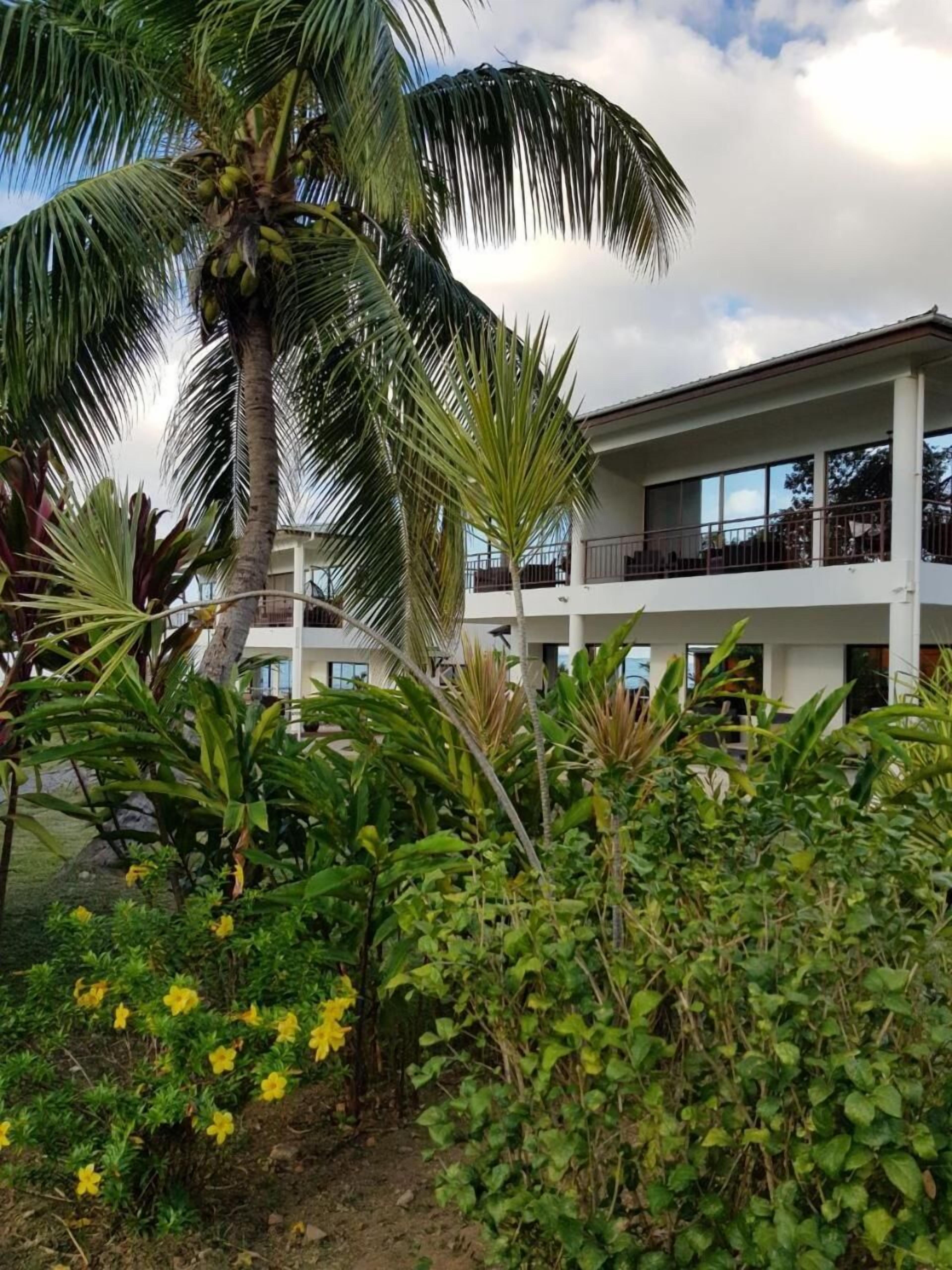 Un edificio de dos pisos con balcones, rodeado de vegetación tropical que incluye palmeras y plantas con flores.