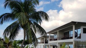 Un edificio de dos pisos con balcón, rodeado de follaje tropical y un cielo despejado.