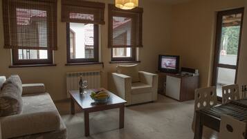 Una sala de estar con un sofá, una mesa con un frutero, un televisor y tres ventanas con persianas.