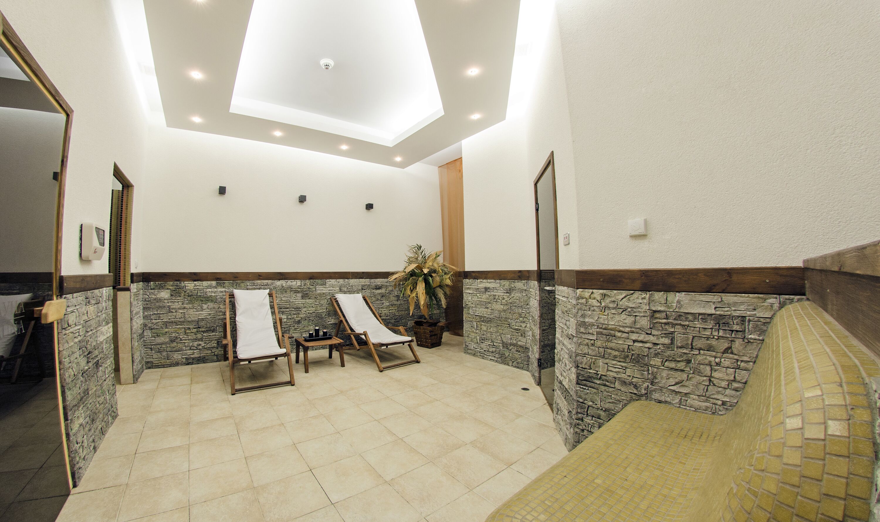 A spa room with two wooden loungers, a stone wall, a ceiling skylight, and a tiled floor.