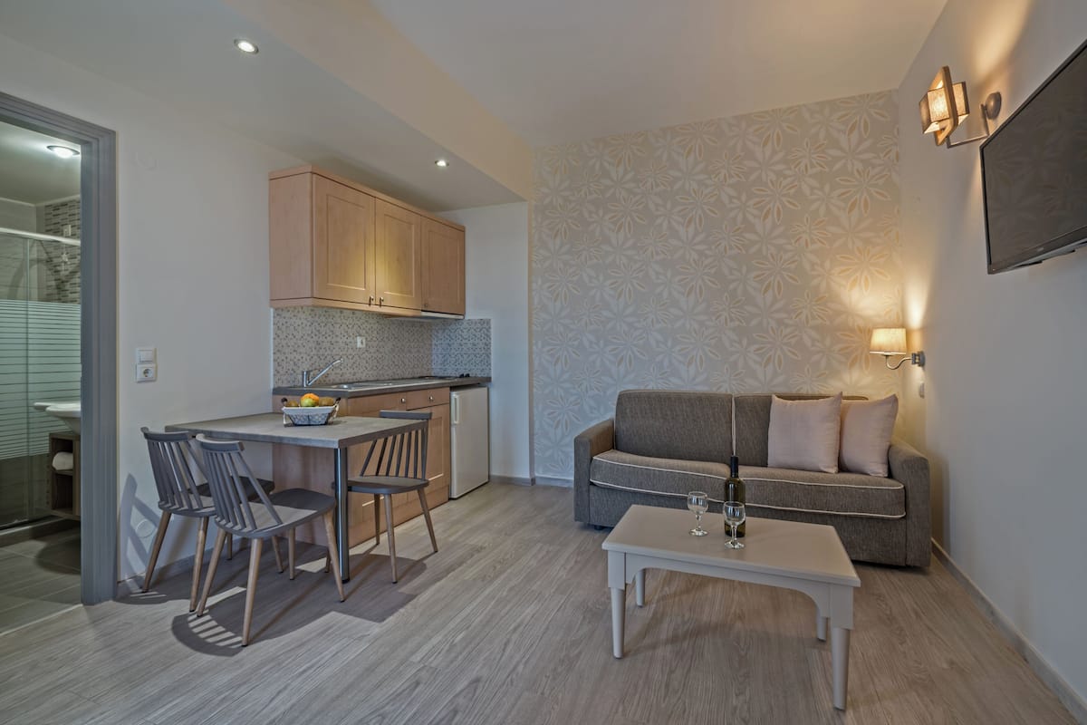 A modern kitchen with wooden cabinets, a dining area with two chairs, a sofa, and a small table with a wine glass.