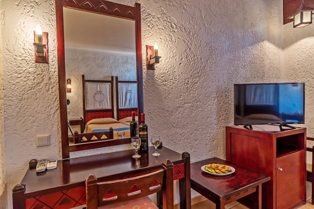 A hotel room with a wooden dining table, chairs, a television on a wooden stand, and a large mirror reflecting the room.