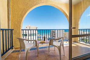 A balcony with a view of the sea, two white chairs, a table with a plate of food, and a bottle of wine.