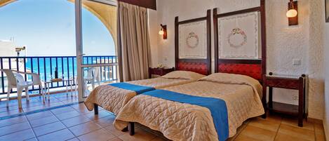 Habitación de hotel con una cama grande, un balcón con vistas al mar y una pequeña mesa con sillas.