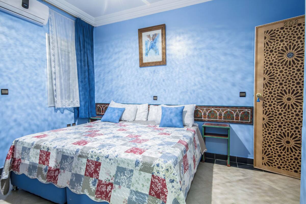 A bedroom with a bed, a nightstand, a wall-mounted air conditioner, a door with a decorative panel, and a framed picture on the wall.