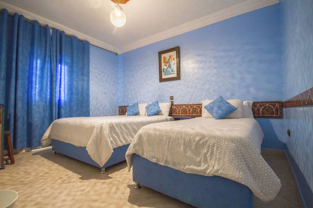 A room with two beds, blue walls, a framed picture, and a light fixture.