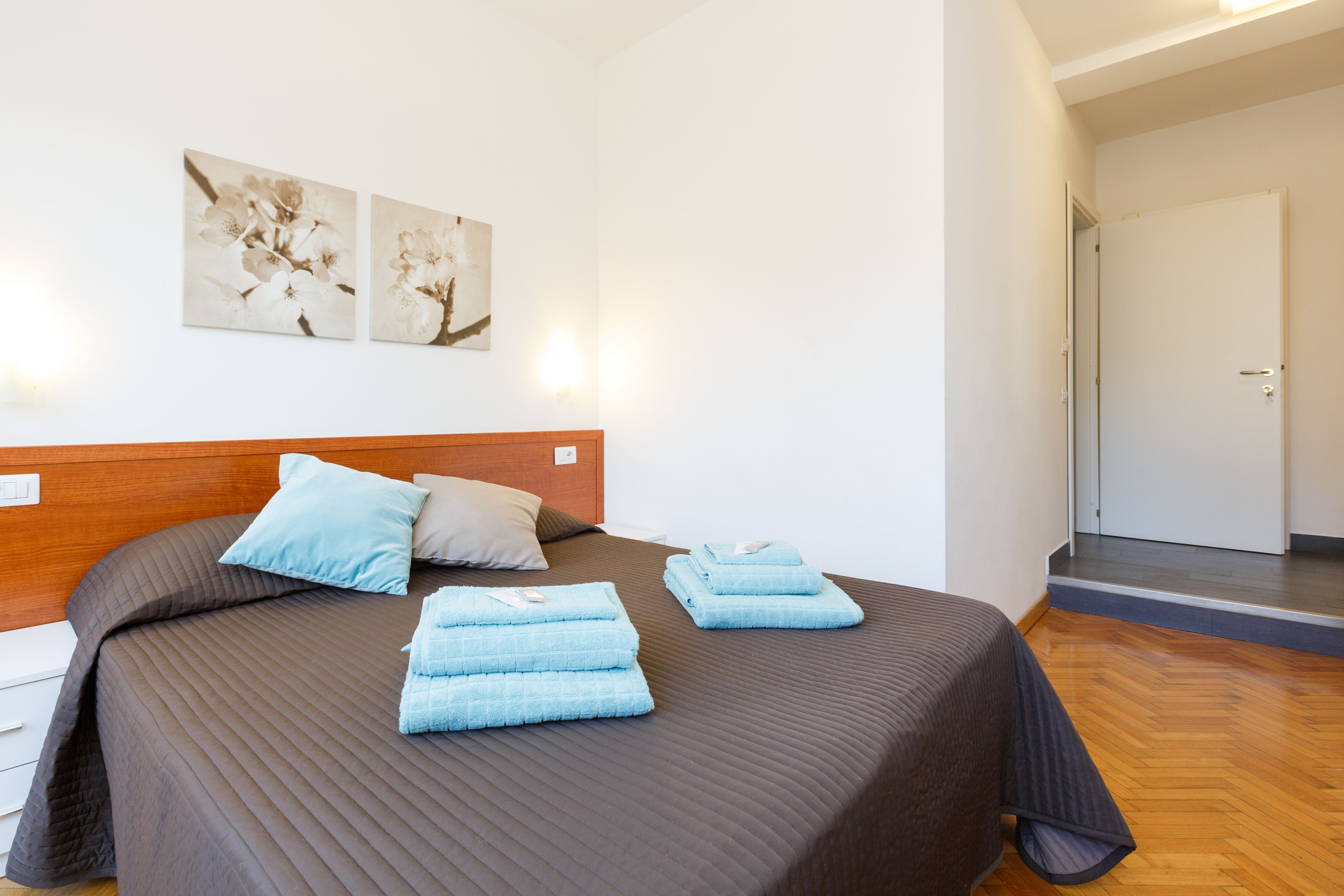Cama bem arrumada com colcha marrom e toalhas azuis, cabeceira de madeira e dois quadros de flores na parede.