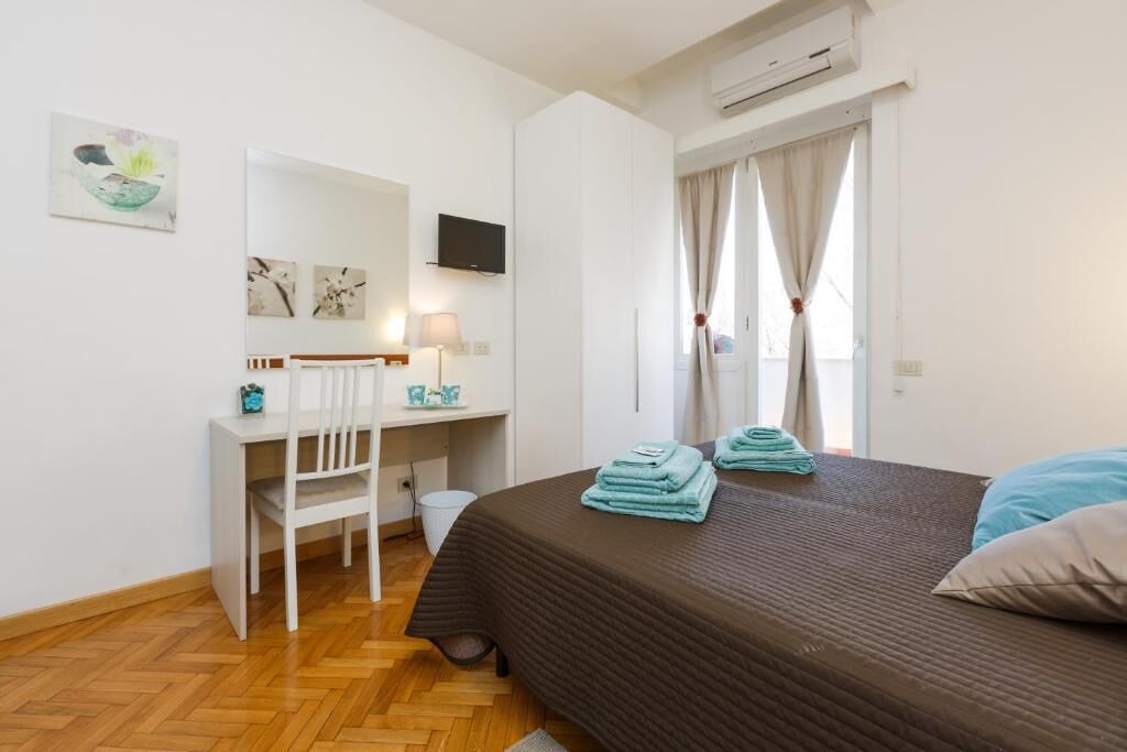 Quarto de hotel com cama, escrivaninha, cadeira, televisão e janela com cortinas.