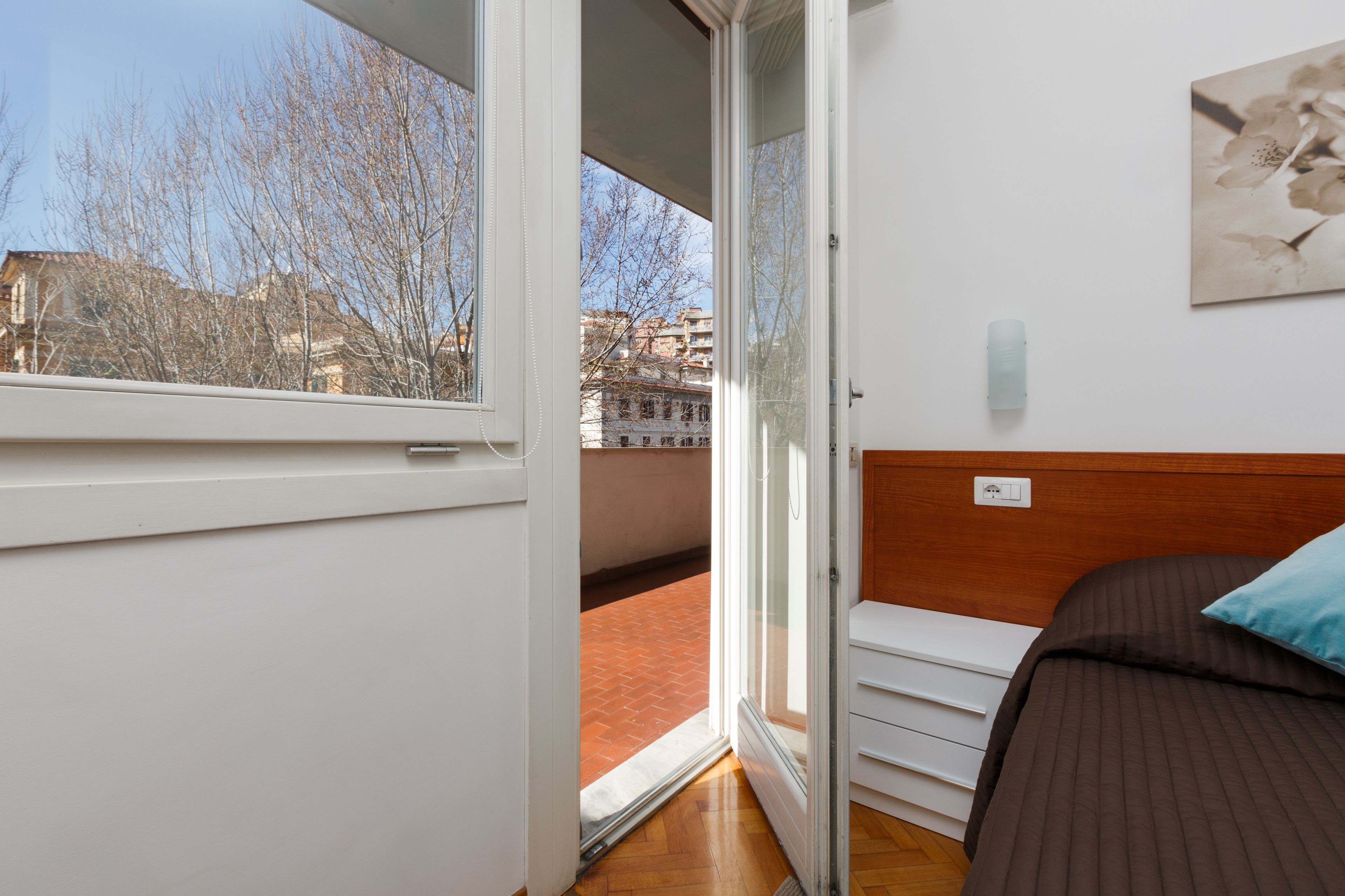 Ein Schlafzimmer mit einem großen Fenster, einem Bett mit brauner Kopfteil und Blick auf Gebäude und Bäume.