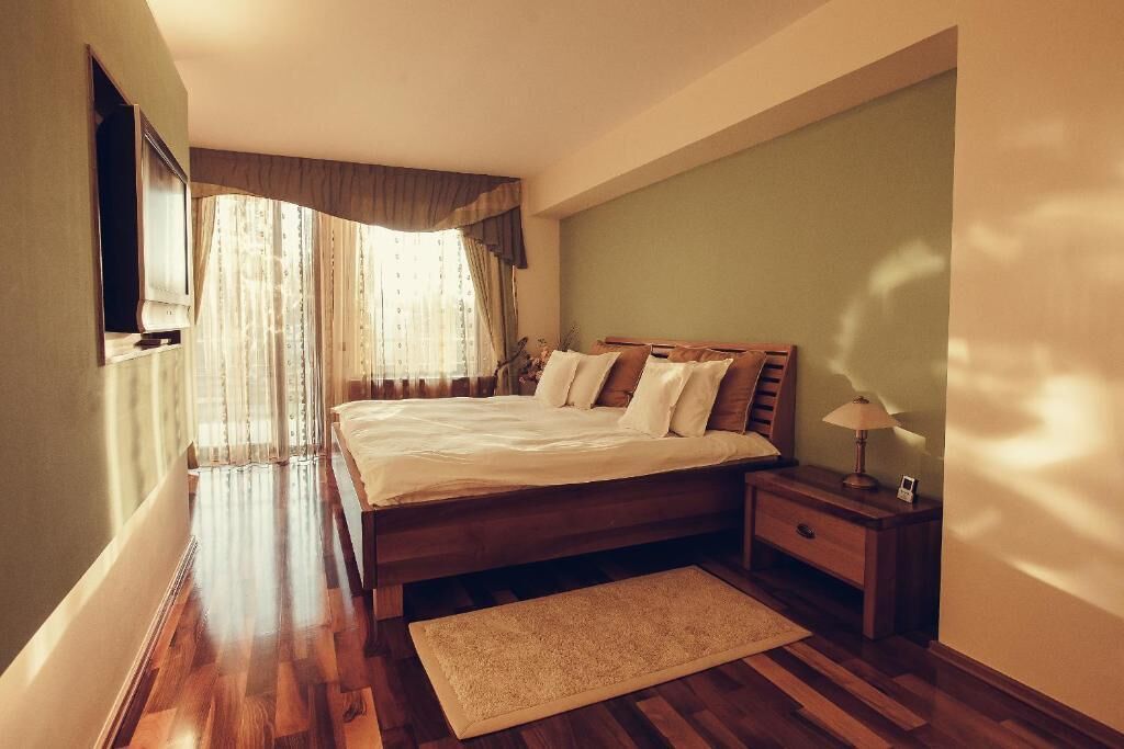 A bedroom with a wooden bed, a nightstand, a lamp, and a large window with curtains.