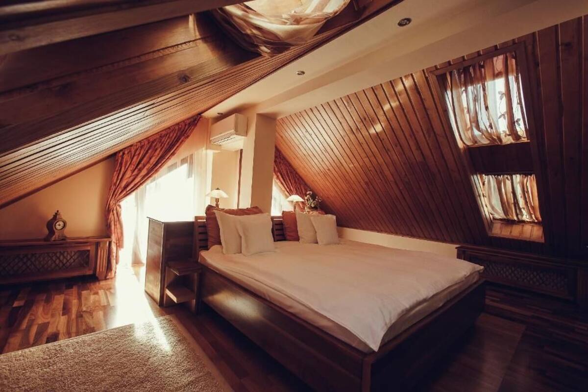 A bedroom with a sloped ceiling, wooden beams, a large bed, a nightstand, and a window with curtains.