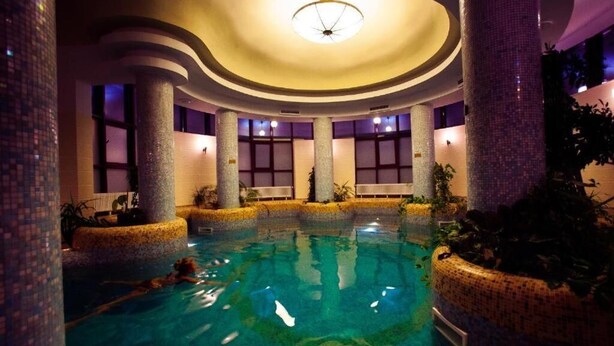 An indoor swimming pool with clear water, surrounded by columns and plants, and a ceiling light fixture. - Denisa Boutique (Brasov)