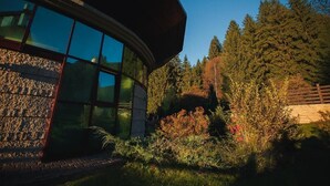 A modern building with large glass windows, surrounded by a garden with various plants and trees. - Denisa Boutique (Brasov)