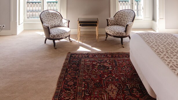 Une pièce spacieuse avec un grand lit, deux fauteuils à motifs, un plafond décoratif et une vue dégagée grâce à des portes ouvertes.