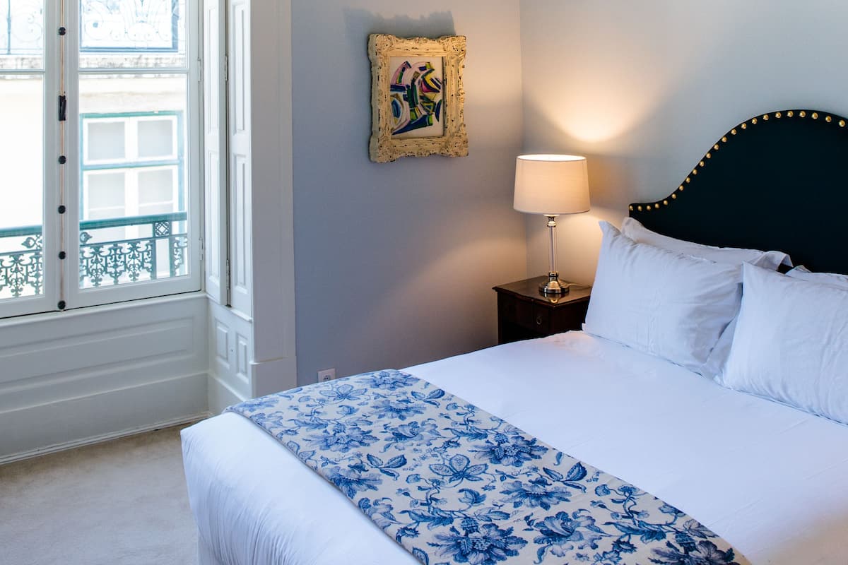 Une chambre à coucher avec un lit, une table de chevet, une lampe, une fenêtre et un tableau encadré au mur.