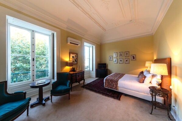 A hotel room with a bed, two chairs, a small table, a TV, and a window with a view of greenery.