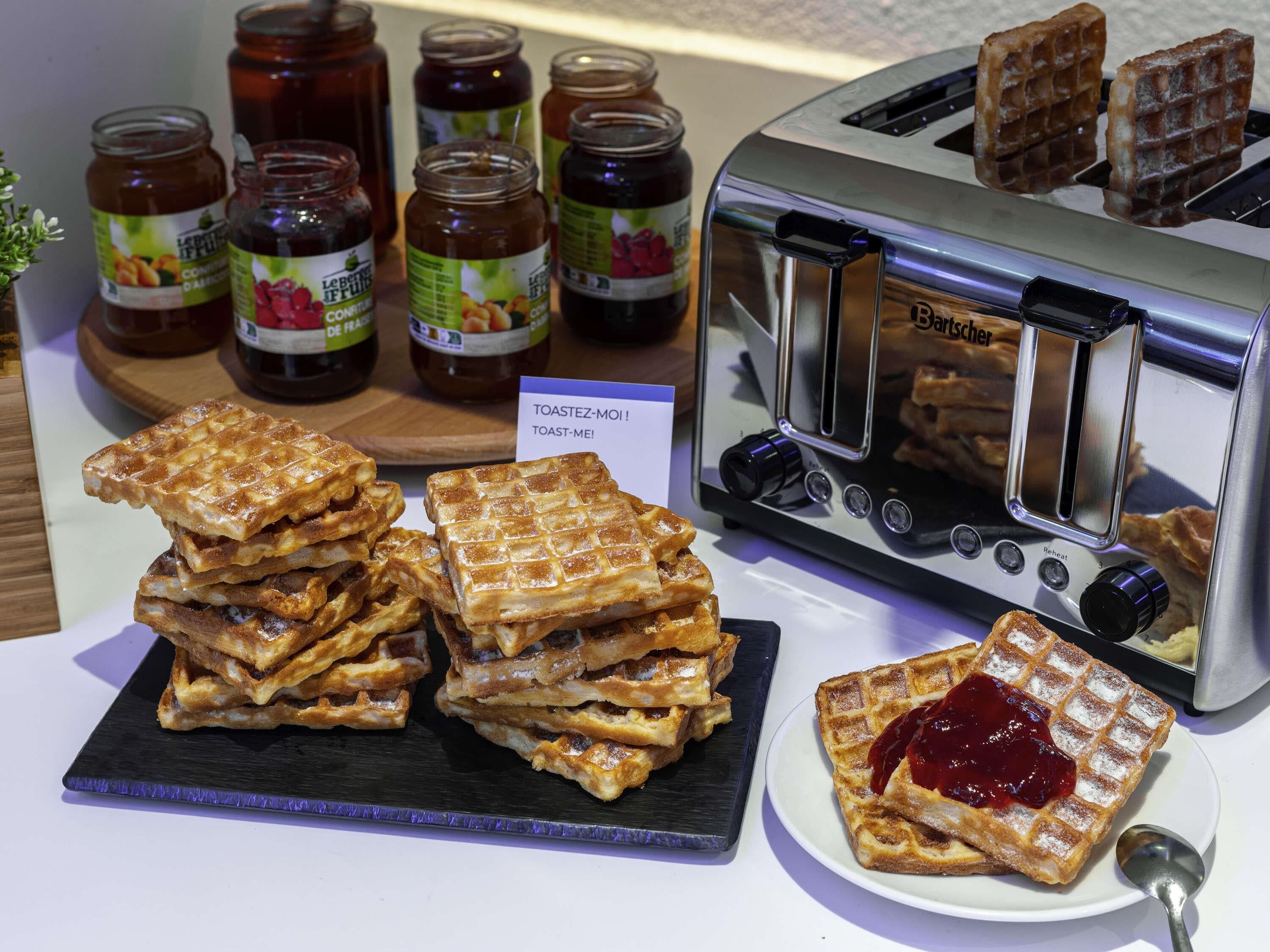 A toaster with waffles, a stack of waffles, and jars of jam.