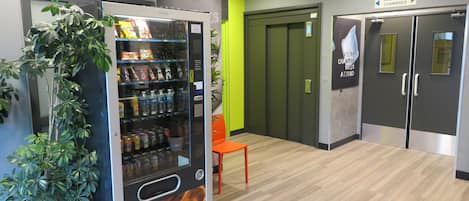A vending machine with snacks and drinks, a green wall, a wooden floor, and a sign for room access.