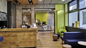 Interior moderno de una cafetería con mostrador de recepción de madera, zona de estar con sillas coloridas y una cocina visible al fondo.