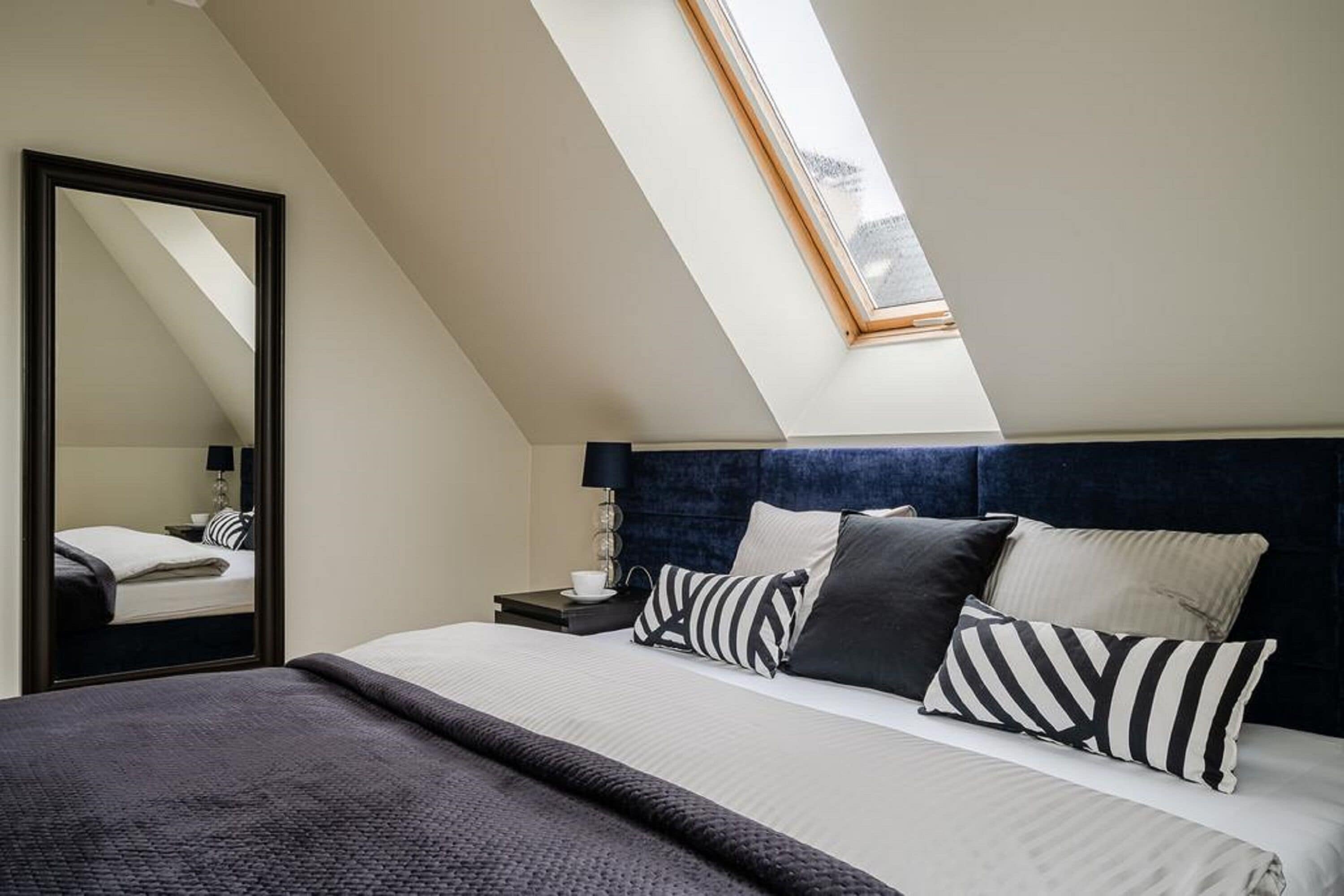 A bedroom with a skylight, a bed with striped pillows, a dark headboard, and a nightstand with a lamp.