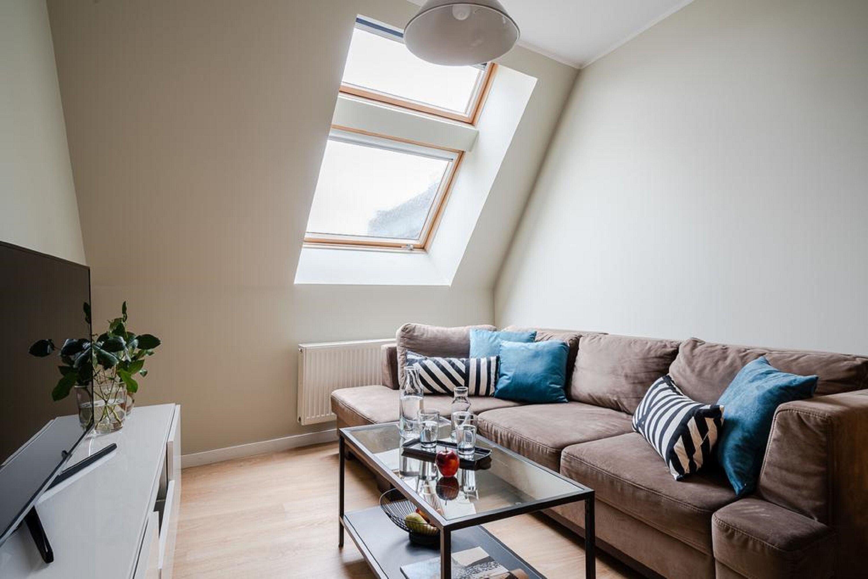 Un salon moderne avec un canapé marron, une table basse en verre et une petite table d’appoint blanche. Il y a deux fenêtres équipées de stores, un radiateur et un luminaire au plafond.