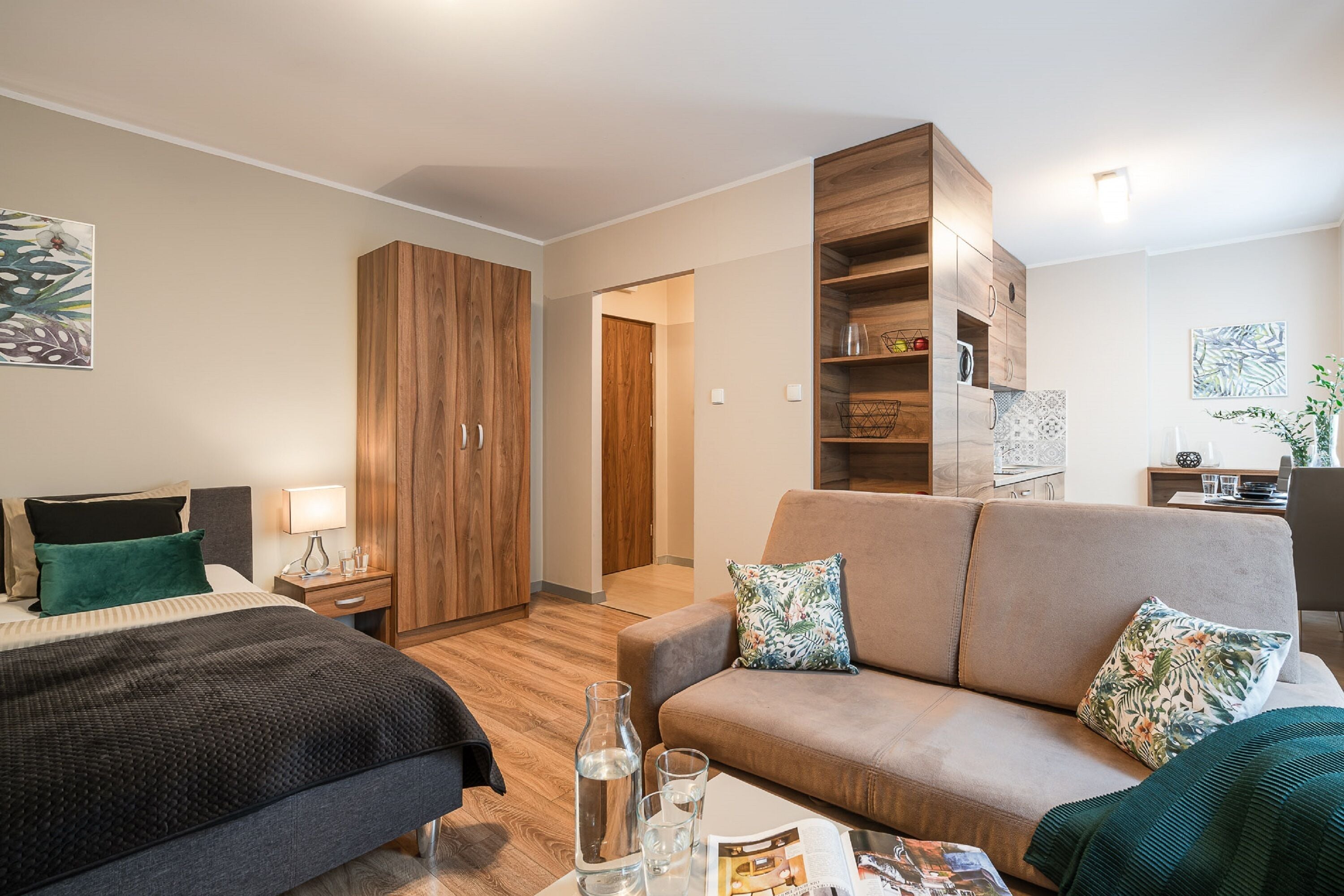A modern hotel room with a bed, a sofa, a wooden wardrobe, and a small table with a magazine and glasses.
