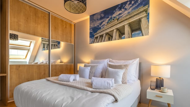 A neatly made bed with white linens and a beige throw blanket, a bedside table with a lamp, and a large painting of a classical building on the wall. - Absynt Apart Wierzbowa (Wroclaw)