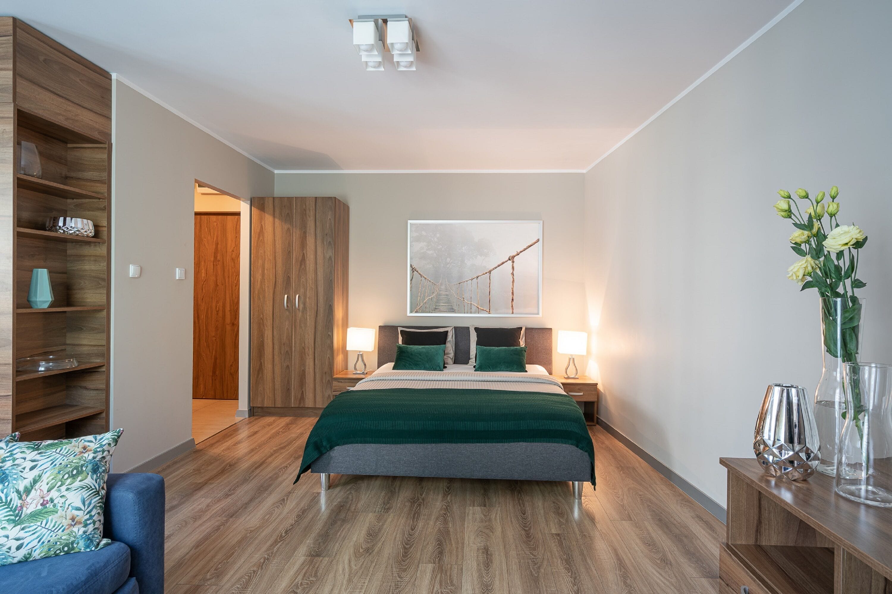 A modern bedroom with a wooden floor, a bed with green bedding, a blue armchair, and a wooden wardrobe.