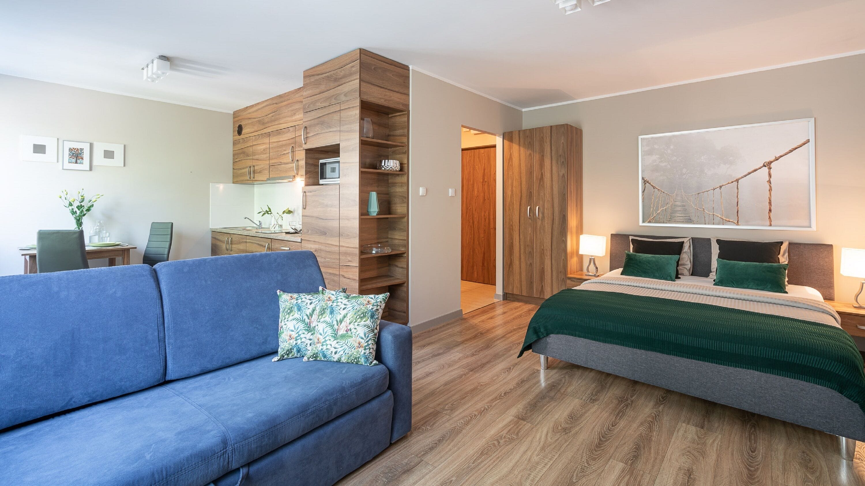 Una habitació d'hotel moderna amb un sofà blau, un llit amb roba de llit verda, panells de fusta i un ampli quadre abstracte a la paret.