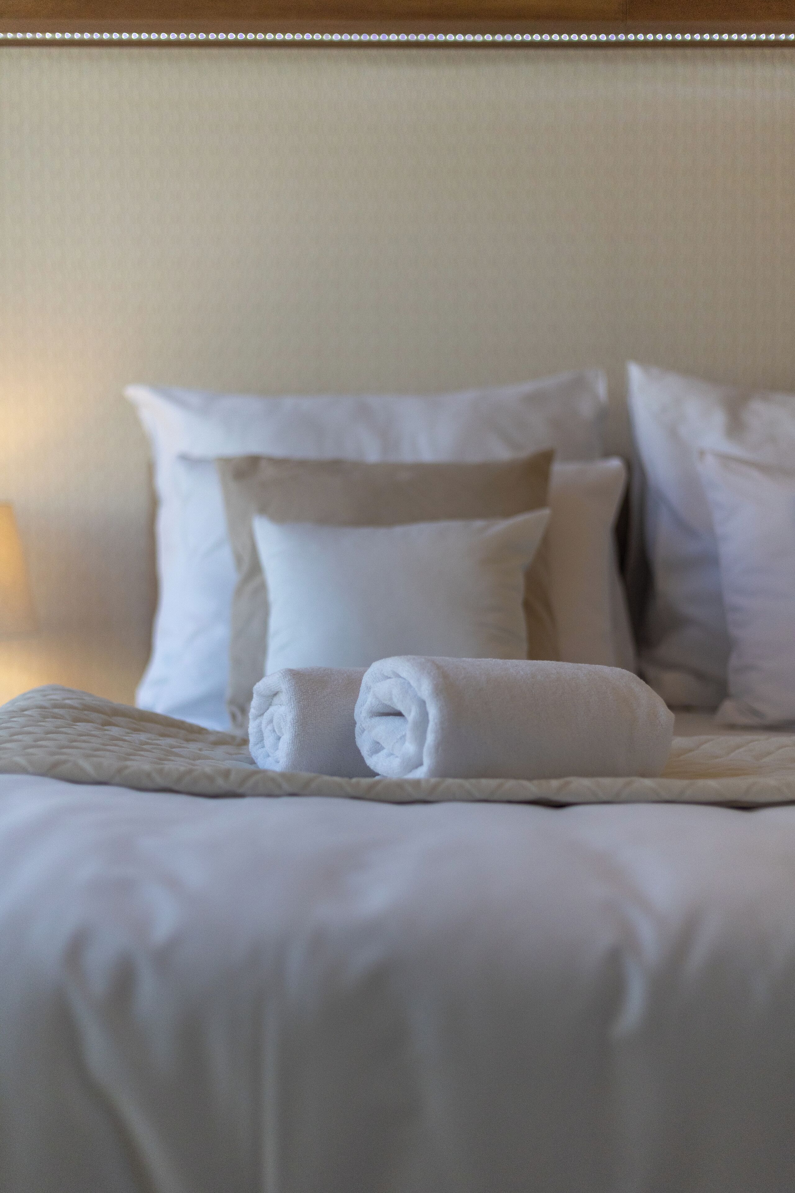 A neatly made bed with white linens and pillows, a rolled towel, and a quilted blanket.