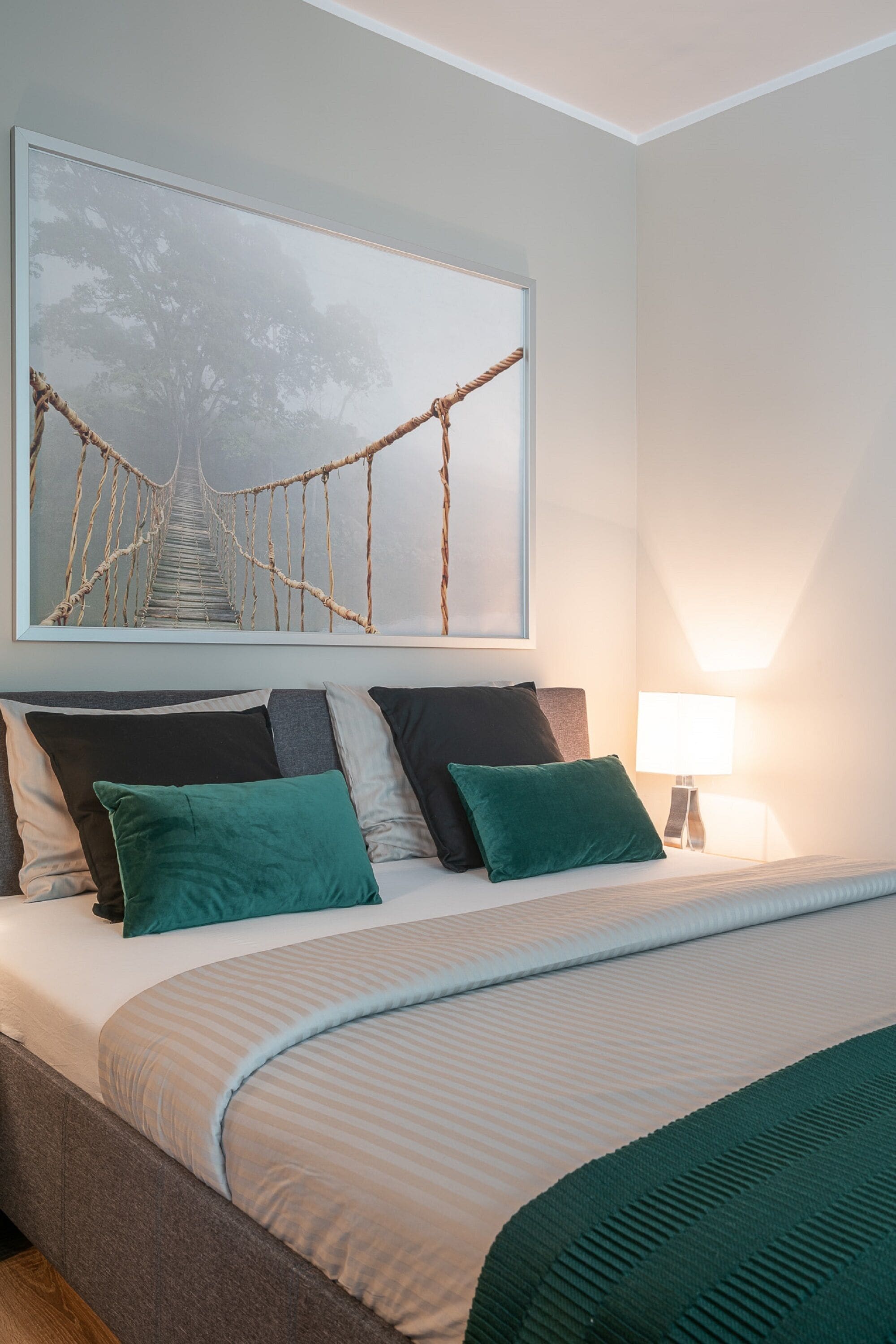 A neatly made bed with a striped bedspread and multiple pillows, a wall-mounted picture of a rope bridge, and a bedside lamp.