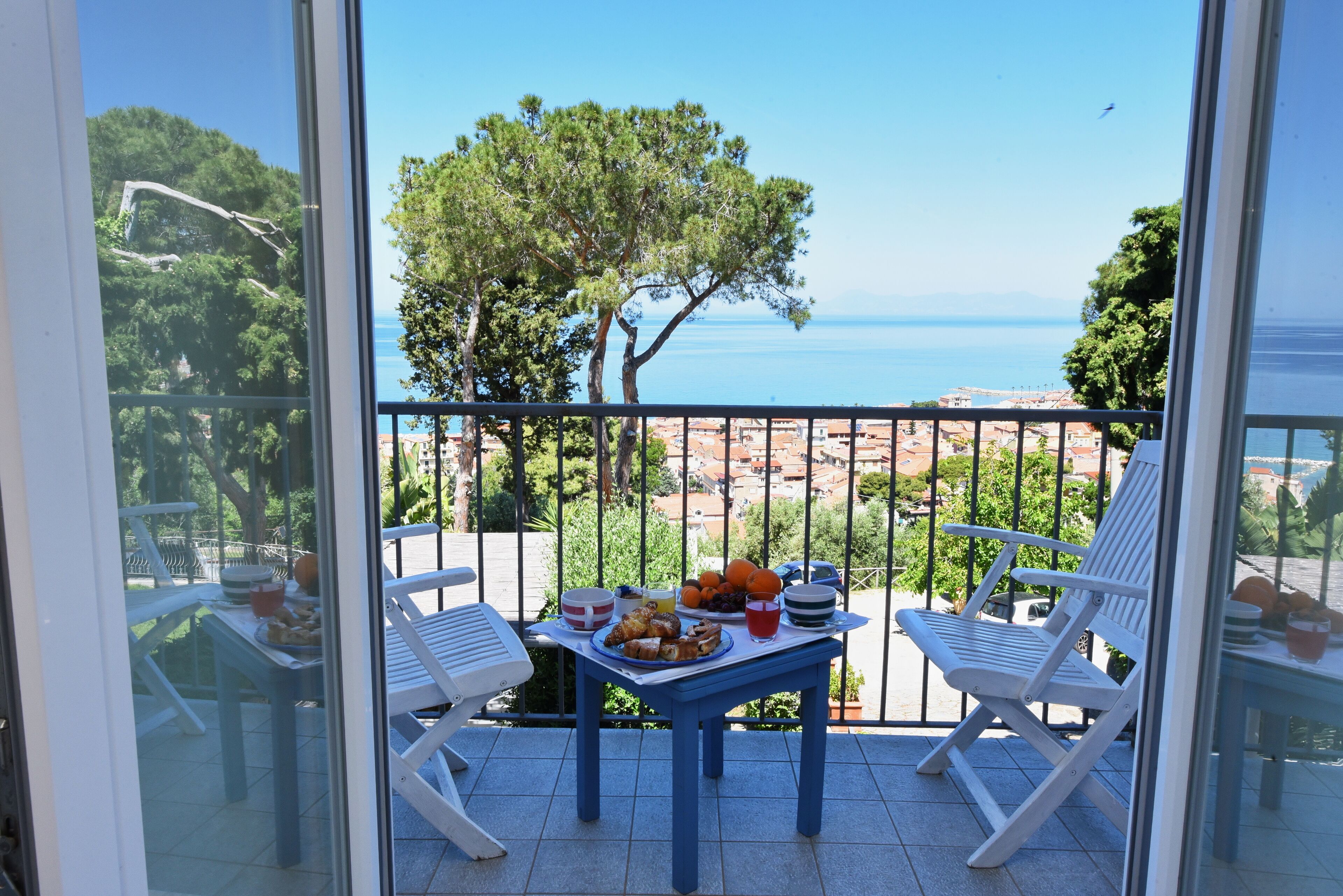 Un balcon avec une table préparée pour le petit-déjeuner, offrant une vue sur la côte, avec des arbres et la mer.