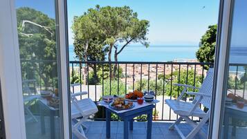 Un balcon avec une table préparée pour le petit-déjeuner, offrant une vue sur la côte, avec des arbres et la mer.