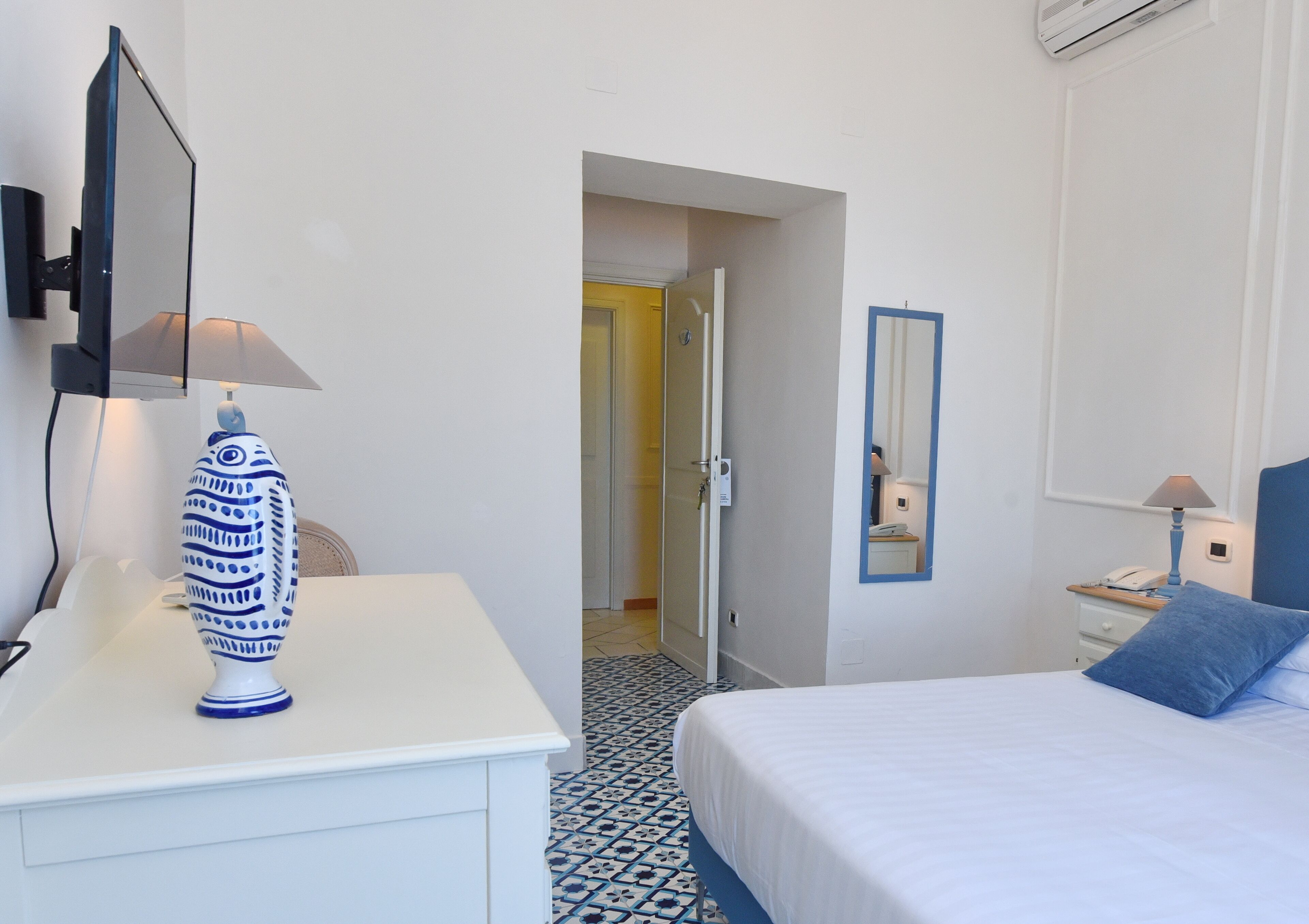 Quarto de hotel com uma cama, uma escrivaninha com abajur, um espelho e uma porta que dá para outro quarto.