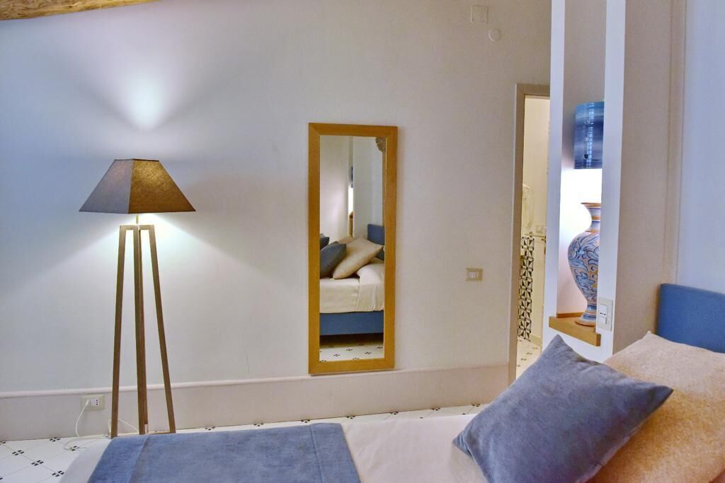 A bedroom with a bed, a bedside lamp, a mirror, and a wall-mounted shelf with a vase.