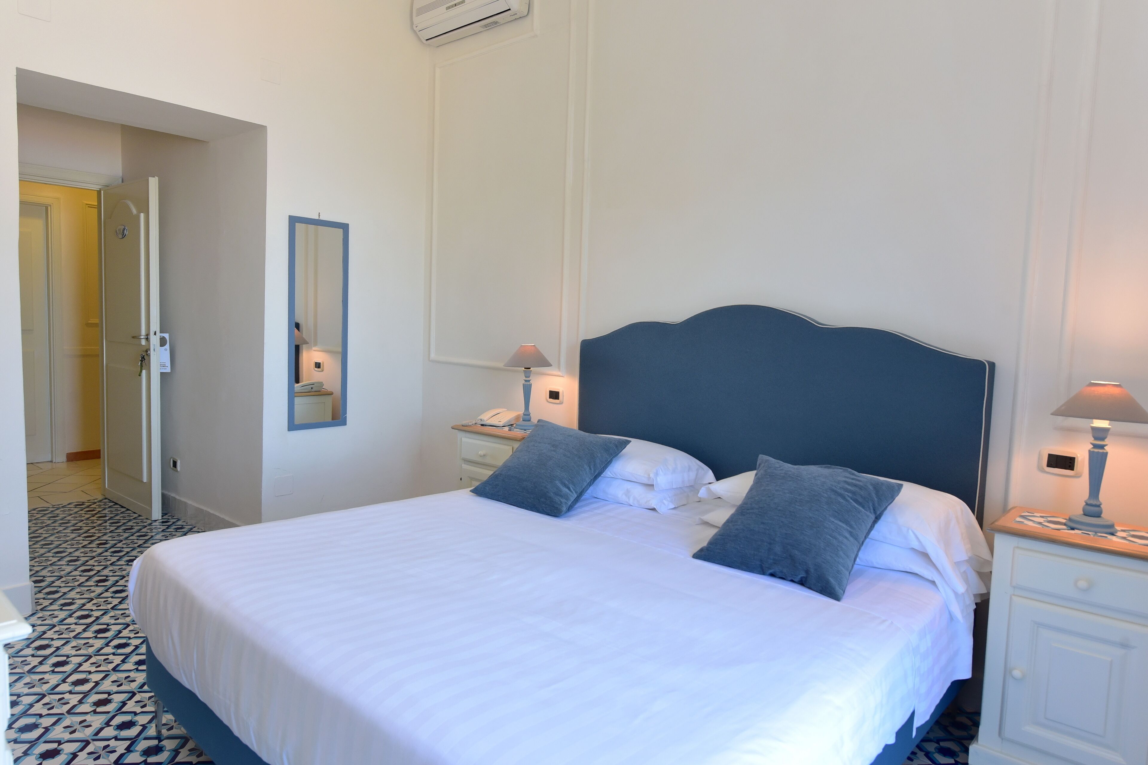 A neatly made bed with white linens and two pillows, a bedside table with a lamp, and a mirror on the wall.
