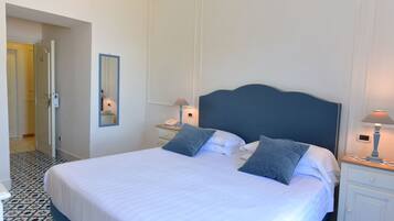 A neatly made bed with white linens and two pillows, a bedside table with a lamp, and a mirror on the wall.