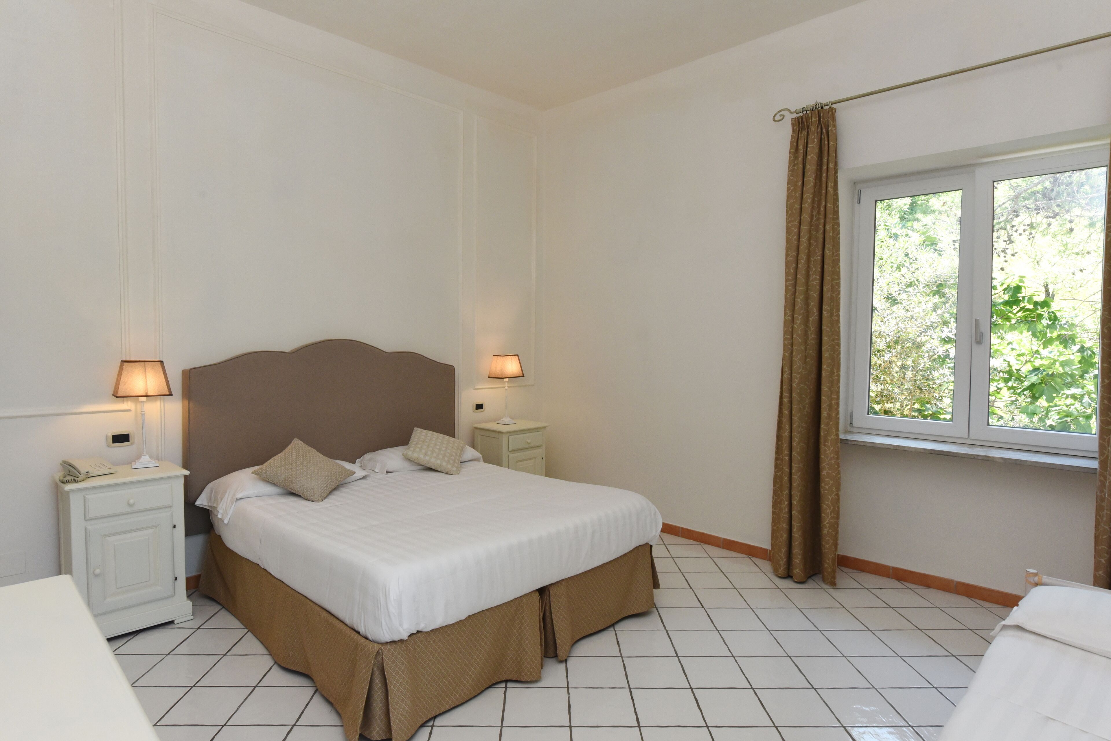 A bedroom with a bed, nightstands, a window with curtains, and a view of greenery outside.
