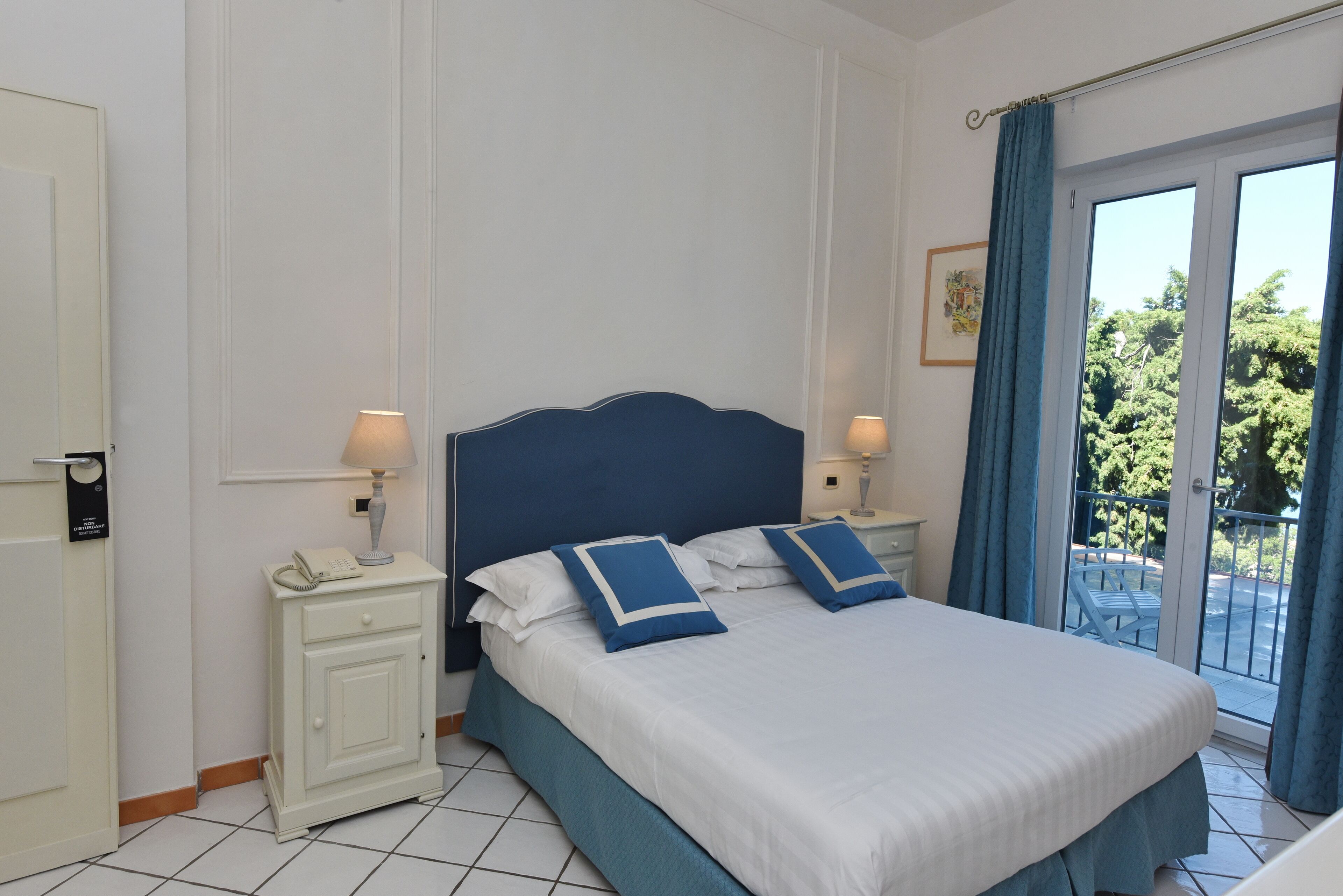 Habitación de hotel con cama, dos mesitas de noche con lámparas, una puerta y vistas al exterior.