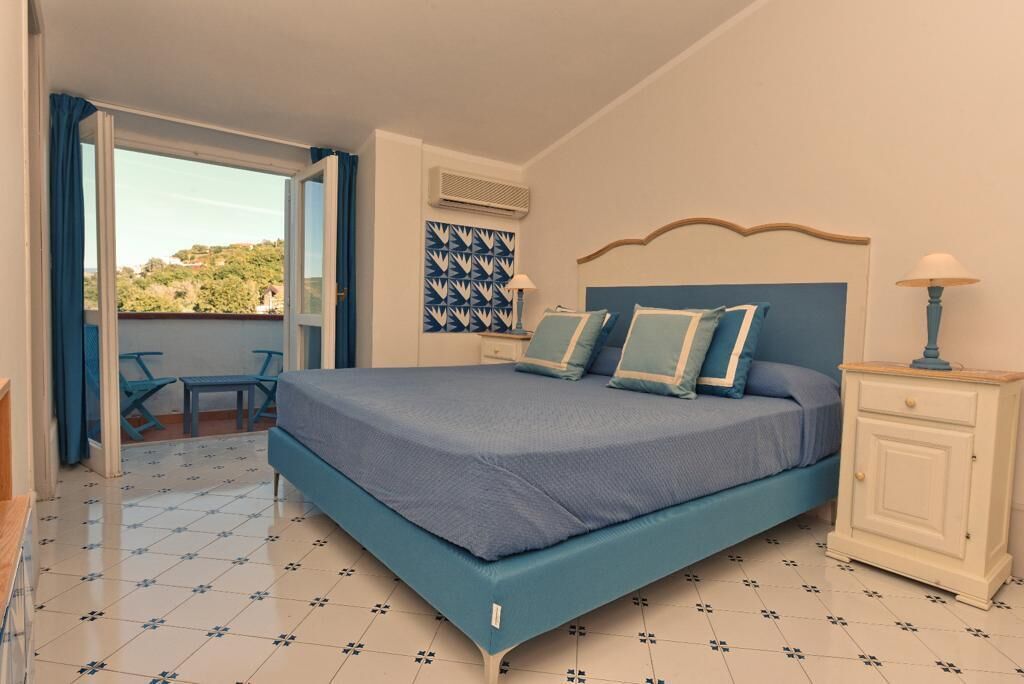 Un dormitorio con cama, mesita de noche, lámpara, pared con estampado, ventana y balcón.