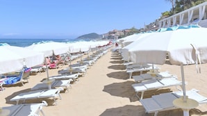 A beach with rows of sun loungers and umbrellas, a clear sky, and a view of the sea and distant hills.
