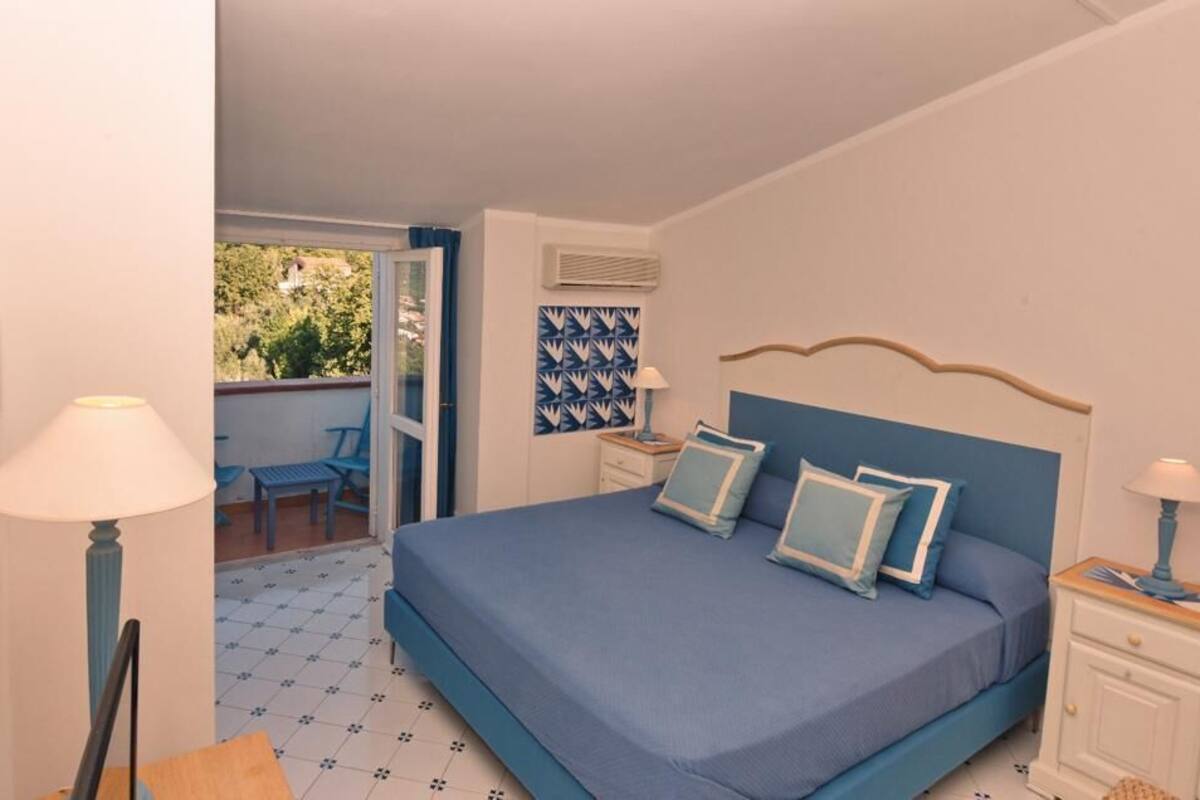 A bedroom with a large bed, a nightstand, a chair, and a view of the outdoors.