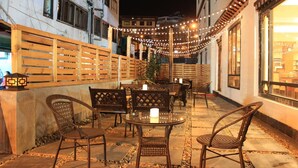 Une terrasse extérieure avec des chaises et des tables en osier, des guirlandes lumineuses et une clôture en bois.