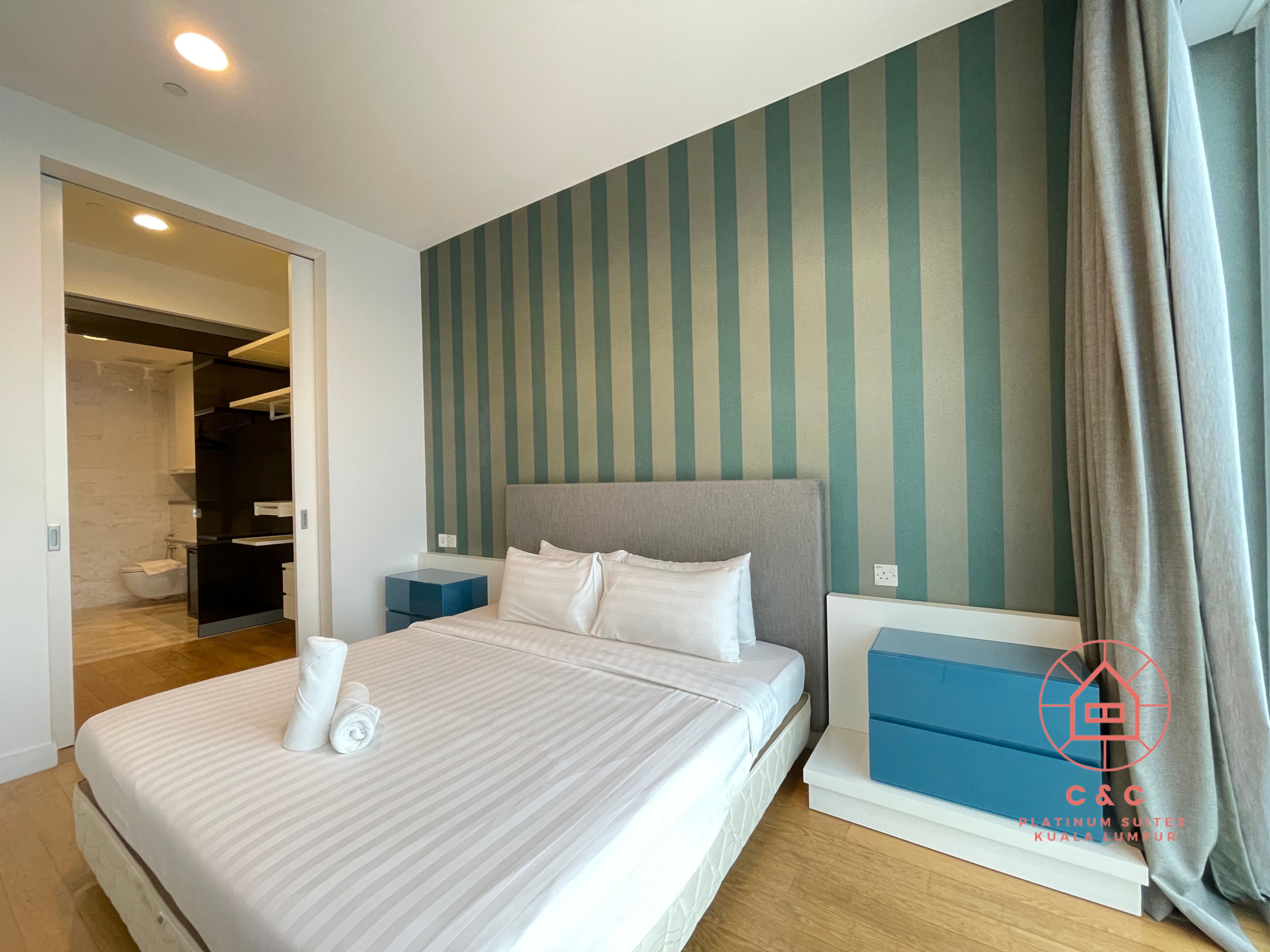 Une chambre d’hôtel moderne avec un grand lit, une table de chevet, une boîte de rangement bleue et une salle de bain visible à travers une porte ouverte.