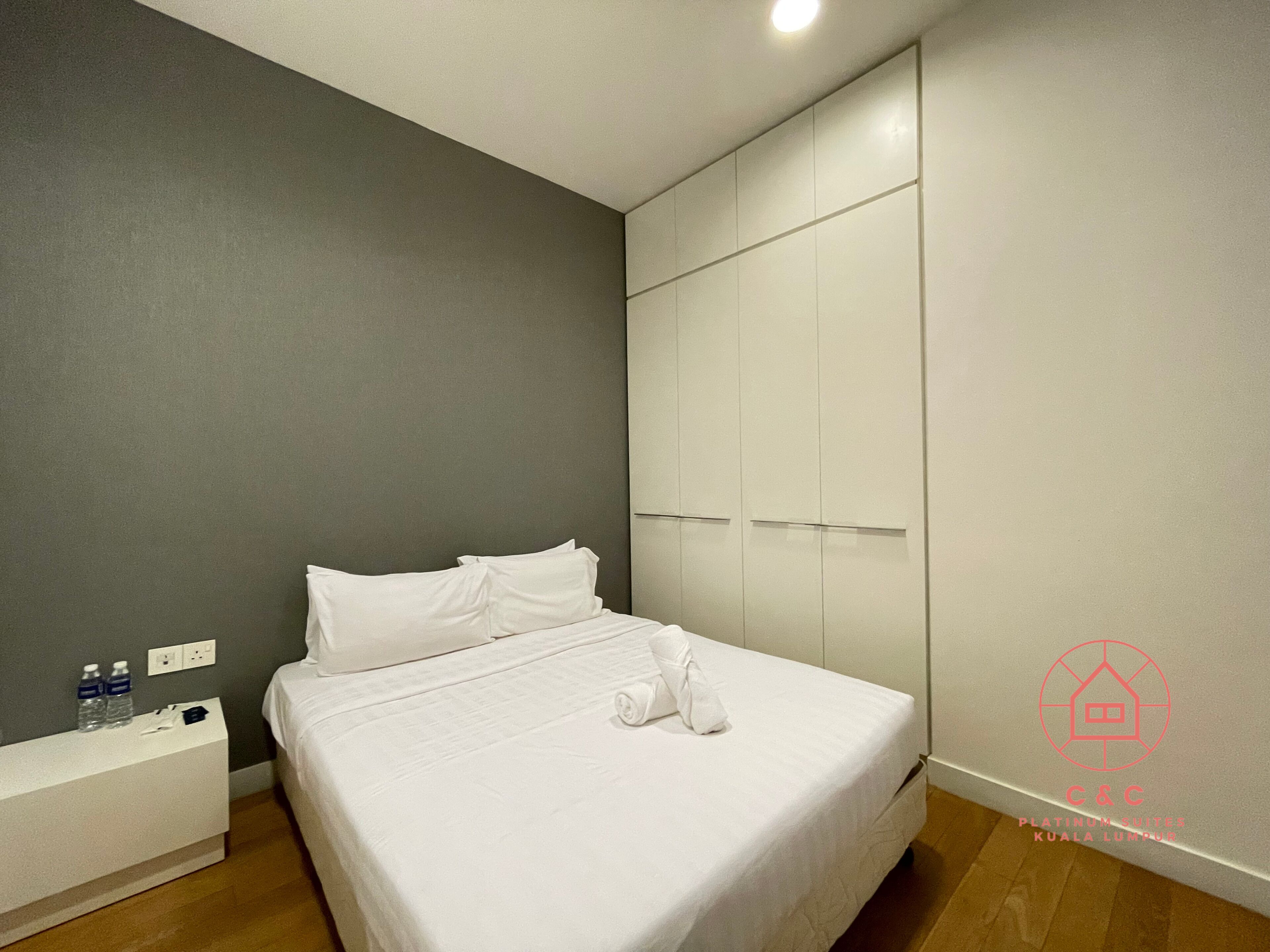 A modern hotel room with a bed, a white bedside table, a wall-mounted wardrobe, and a wooden floor.
