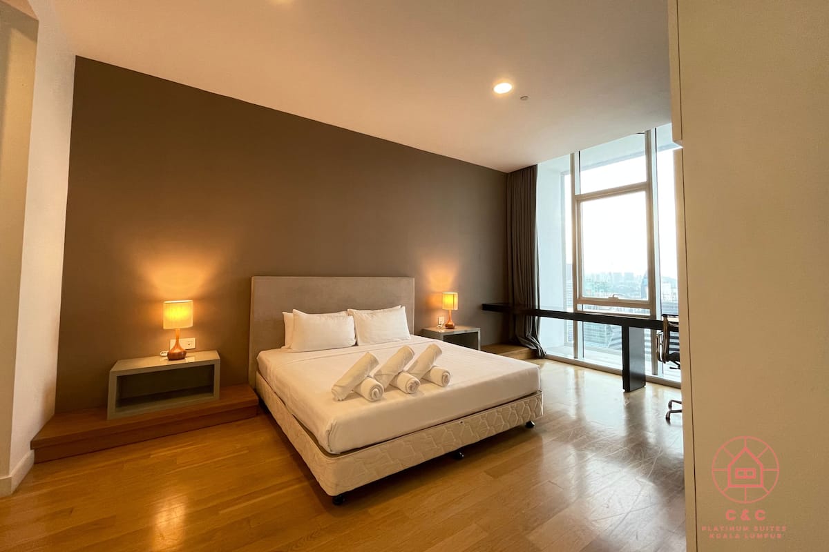 A modern hotel room with a large bed, two bedside tables with lamps, a view of the city through large windows, and a wooden floor.