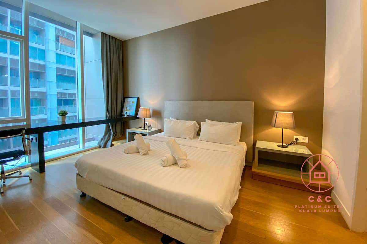 A hotel room with a large bed, two bedside tables with lamps, a desk with a chair, a large window with curtains, and a view of a building.