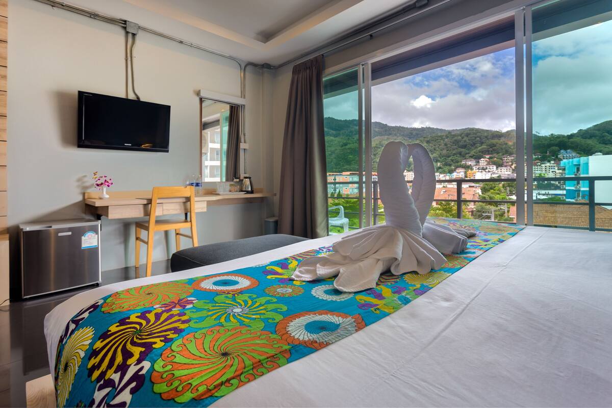 A hotel room with a bed, a desk, a TV, and a view of a city and mountains.
