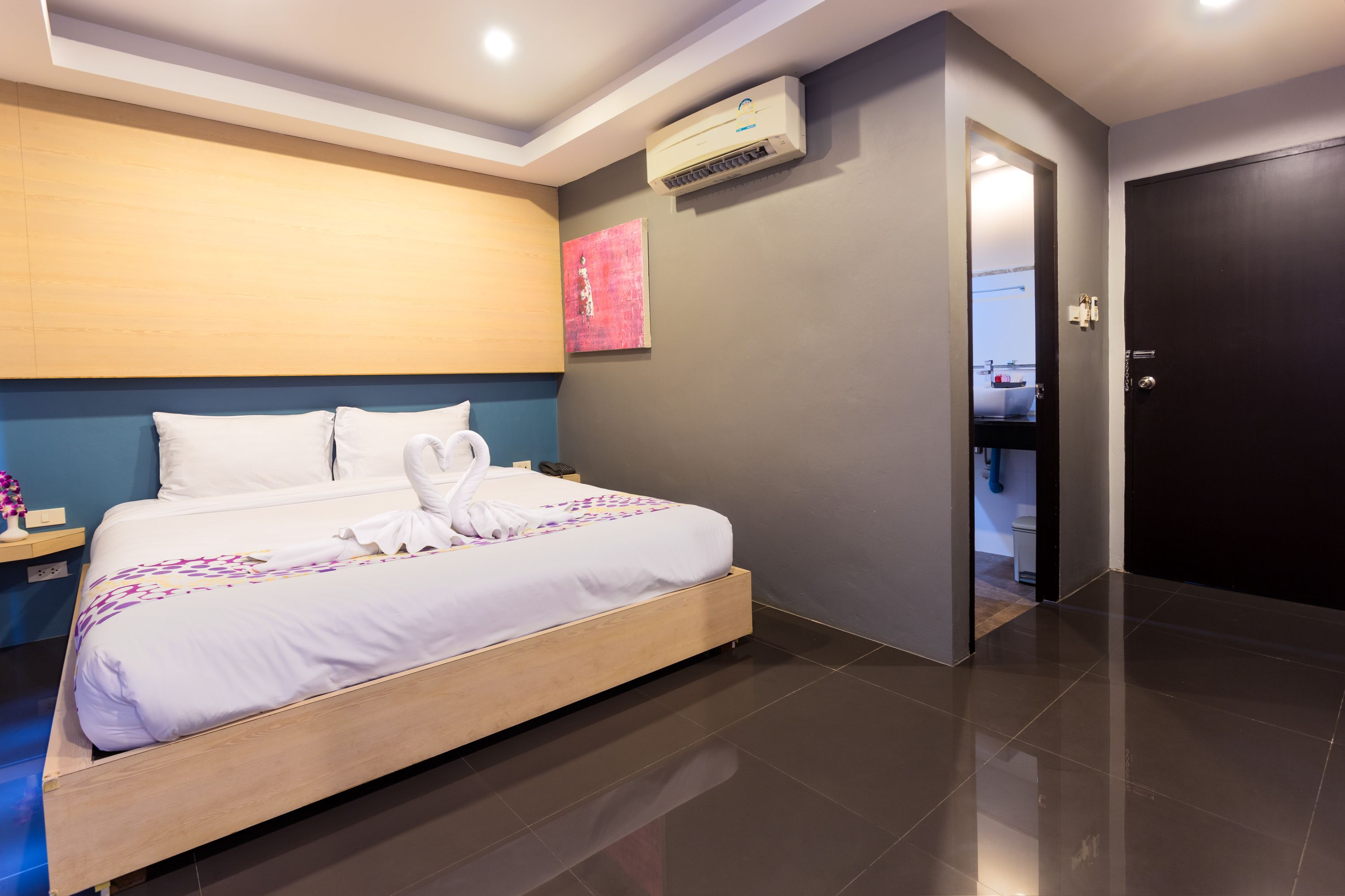 A modern hotel room with a large bed, a wooden headboard, an air conditioning unit, and a door leading to a bathroom.